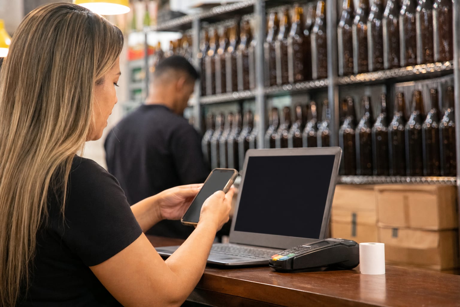 Mãos com celular e notebook no balcão de recebimento de uma distribuidora, com prateleiras ao fundo
